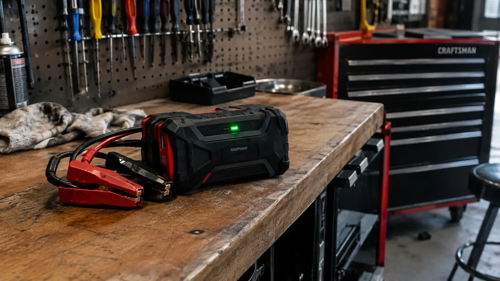 A jump starter device with a charge indicator light glowing green, sitting on a workbench next to a rolling tool chest